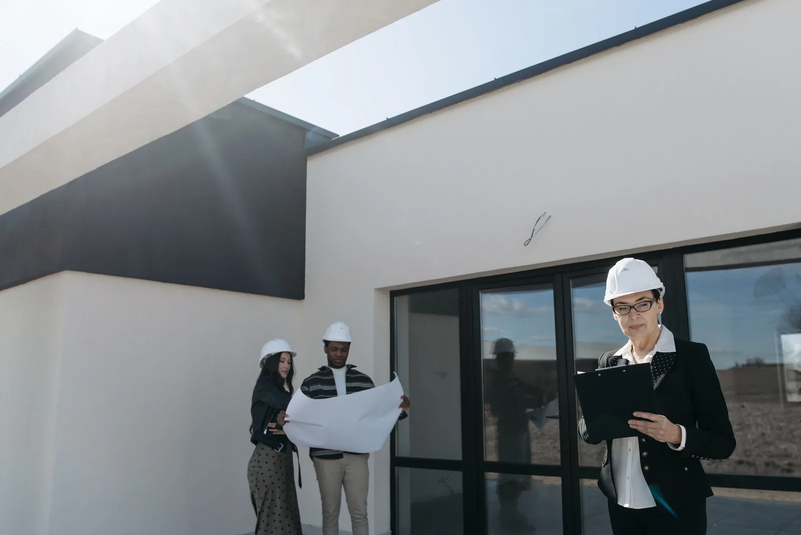 Engenheira com capacete e prancheta conduzindo inspeção técnica de uma edificação, com a equipe analisando a planta ao fundo.